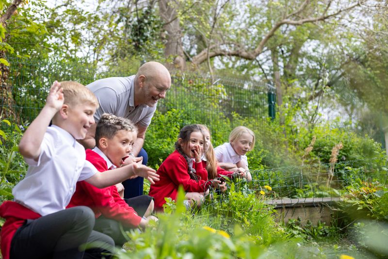 A wide shot of a group of school children sitting around a pond outdoors with their male teacher. They are all learning about the environment and pond wildlife. The children are dressed in red school uniforms with white shirts and black pants. Videos are available similar to this scenario.