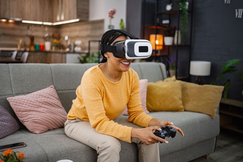 Smiling mid adult woman using virtual reality headset and controller while playing video games at home