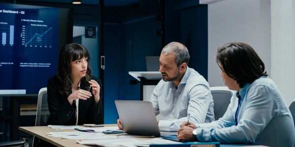 Three professionals engaged in a business meeting with charts displayed on a screen.