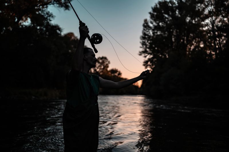 A serene scene featuring a fisher silhouetted against a vibrant sunset, casting a fly rod in calm waters, surrounded by lush trees. Reflects tranquility, skill, and the love for outdoor activities.