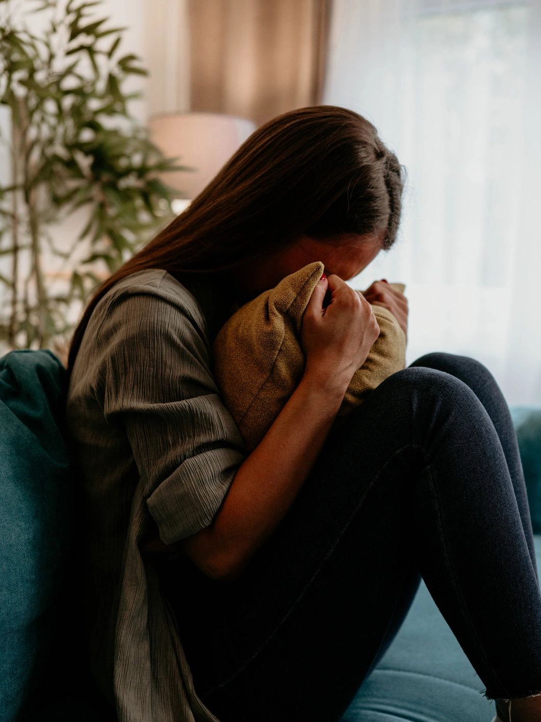 A woman sits curled up on a couch, clutching a pillow and hiding her face.