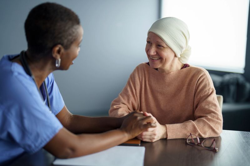 A nurse offers comfort to a cancer patient wearing a headscarf, creating a moment of empathy and support in a clinical setting. The imagery evokes compassion, care, and the human touch in healthcare.