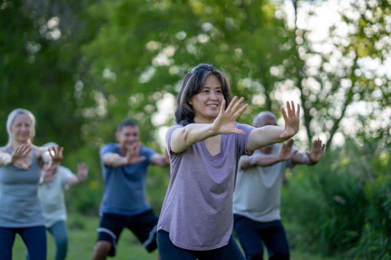 A small group of mature adults practice Tai Chi outdoors together on a warm summer day.  They are each dressed comfortably and are smiling as they take in the fresh air.