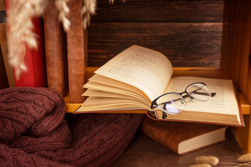 Cozy comfortable nook at library with open hardcover book, warm autumn sweater, dried plants and eyeglasses prepared for evening leisure literature reading and relaxation or hygge knowledge studying