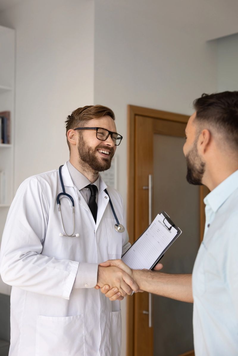 Positive young Caucasian doctor man wearing lab coat and glasses, shaking hands with patient, laughing, holding clipboard, wishing to get better after consultation, successful treatment