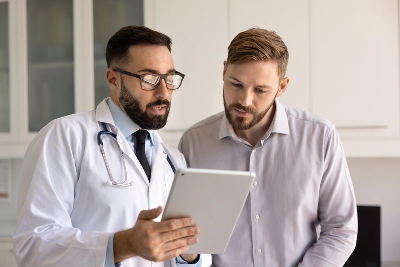 Serious Hispanic medical practitioner man and young patient looking at electronic medical report, examination results on tablet. Doctor explaining diagnosis, online prescription to man