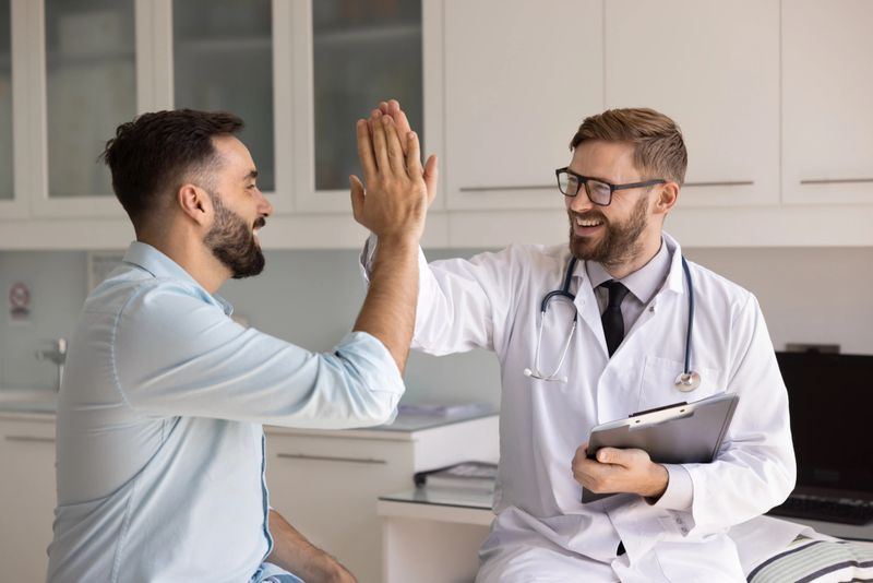 Happy excited young male doctor and healthy patient giving hand high five gesture, smiling, laughing, sitting on medical couch in exam room, celebrating successful therapy over