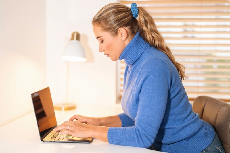 A woman in a blue sweater sits at a desk typing on a laptop, surrounded by a clean and bright workspace with soft natural light coming through wooden blinds.