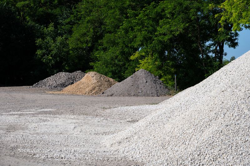 Multiple piles of gravel and crushed stone are neatly arranged on a depot. The image shows various textures and colors of the materials, set against a backdrop of green trees and a clear blue sky.