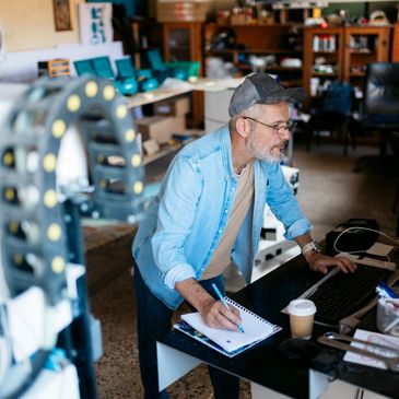 Man working at a cluttered desk, taking notes while using a computer.
