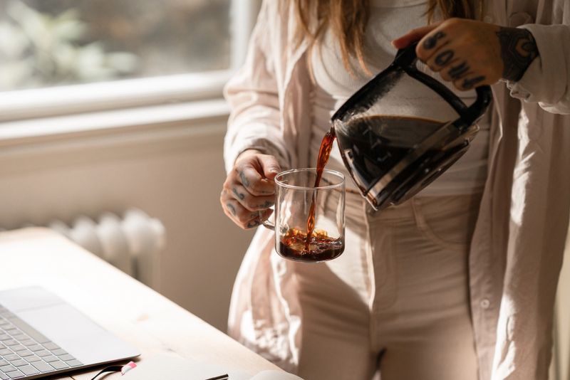 A woman with tattoos pours coffee into a clear mug by her workspace. Sunlight brightens the peaceful scene, evoking calm and focus during her home-office routine.