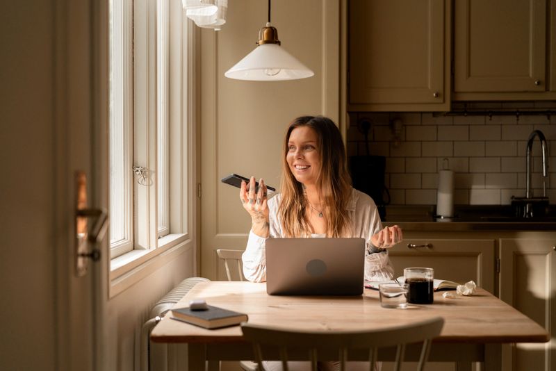 Focused woman working from home at her kitchen table, using a smartphone for voice commands. The laptop and notebook suggest she's multitasking efficiently in a comfortable, sunlit interior environment.