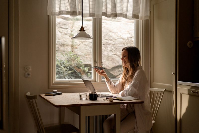 A woman holding her smartphone and interacting with an AI assistant while working at a cozy home space. The ambiance is calm, showcasing a wooden table by a sunny window.