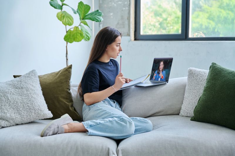 Teenage girl college student having online lesson, watching webinar, sitting at home on couch using laptop computer. Video technology education e-learning distant study knowledge adolescence concept