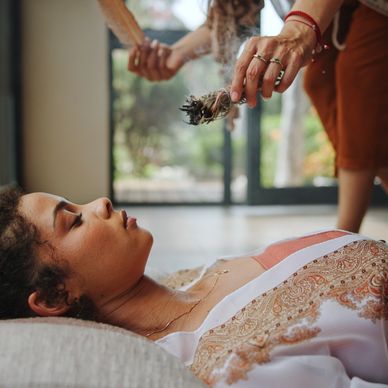 A woman lies down while another person holds smudge sticks above her for cleansing.