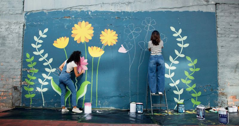Women, creative or friends painting on wall for mural, drawing flowers or community service at building. Artist, outdoor or girls with teamwork for floral decoration, renovation or project for design