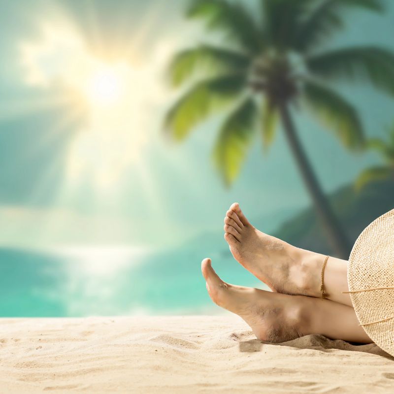 Close-up of a woman’s bare feet gently resting on warm beach sand, with soft sunlight and a clear blue sky in the background. The image captures a peaceful summer moment, evoking feelings of freedom, rest, and sun-soaked relaxation. With space for product placement, it’s perfect for promoting skincare, travel destinations, beachwear, or lifestyle brands focused on self-care and summer vibes.