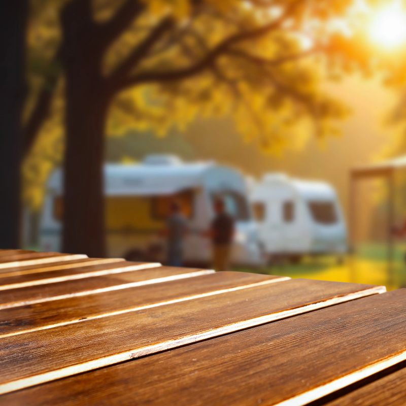 Close-up of a wooden table with empty space in the foreground, perfect for product placement or branding. In the blurred background, camping trailers and tall trees evoke a peaceful outdoor lifestyle and a travel-friendly atmosphere. This image is ideal for showcasing camping gear, food, drink, or nature-inspired products in a casual and natural setting.
