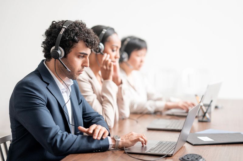 A group of professionals working in a call center, wearing headsets, focused on delivering quality customer support. They are seated at a desk using laptops in a modern business setting.