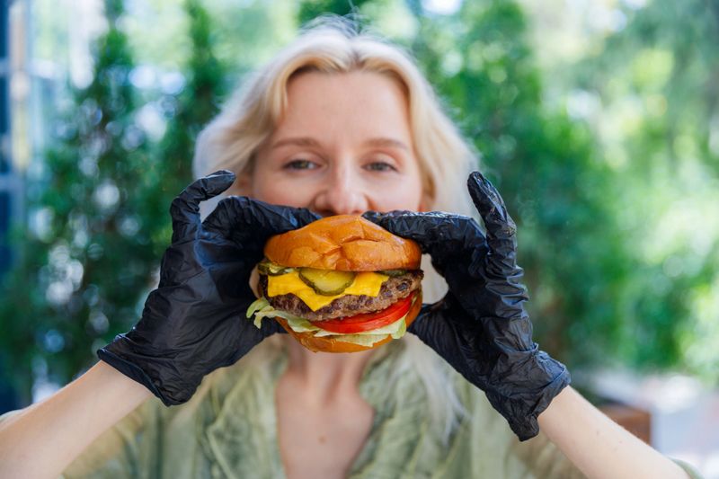 A cheerful person wearing black gloves proudly holds a juicy cheeseburger topped with lettuce, tomato, and pickles in front of their face, set against a lush green background.