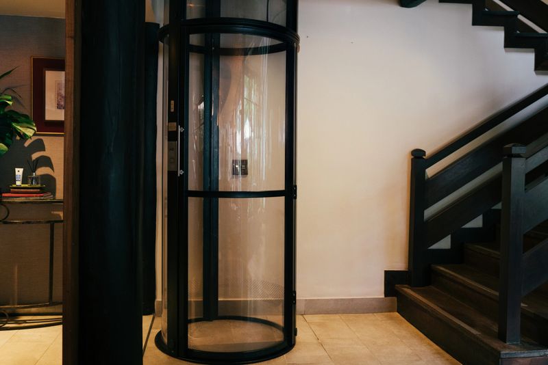 A cylindrical glass elevator providing vertical access within a modern home, positioned next to a wooden staircase.