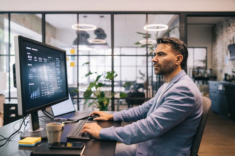 A professional adult male worker reviews data on a desktop monitor in a modern office environment, wearing smart casual business attire, with focused expression and strategic planning concepts.
