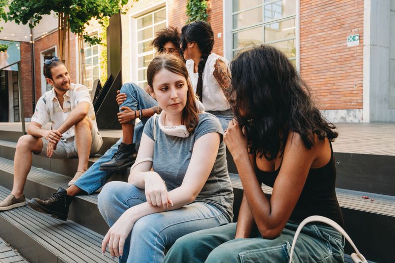 Group of young adults engaging in conversation while sitting on steps in a modern urban setting. Casual and relaxed atmosphere, highlighting friendship and connection