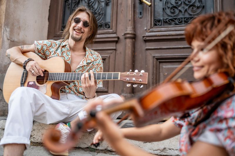 Outdoor scene of a guitar player with a friend playing violin, sharing music and smiles.