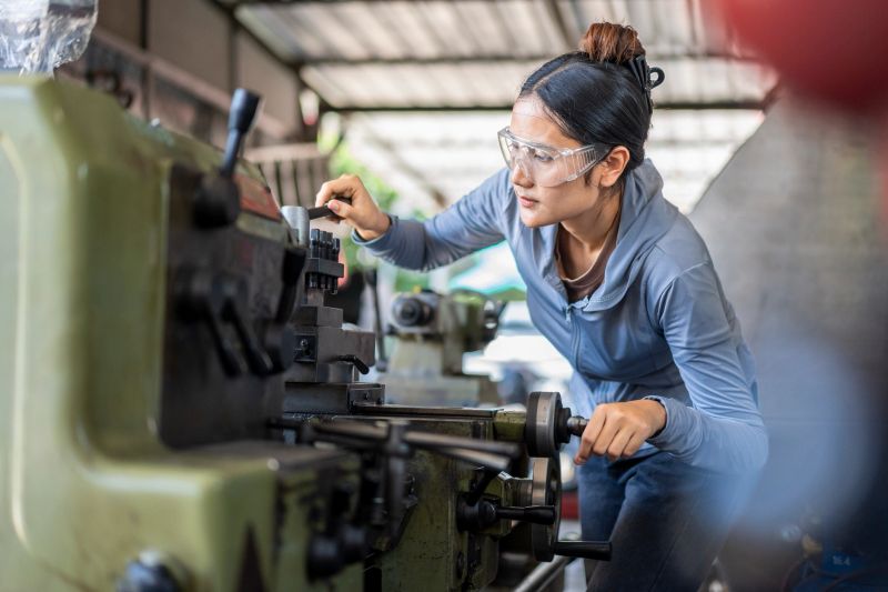 A skilled Asian woman machinist wearing safety glasses meticulously works on a metal lathe machine in a busy industrial workshop, focusing on precision and craftsmanship.