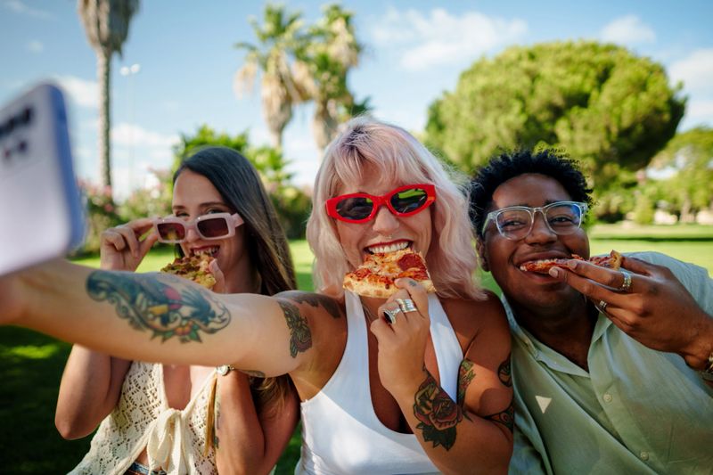 Three cheerful young friends are taking a selfie while enjoying pizza in a park