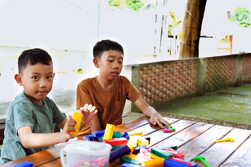Two boys crafting together using colorful paper and tools at a picnic table outside. Educational and hands-on learning moment.