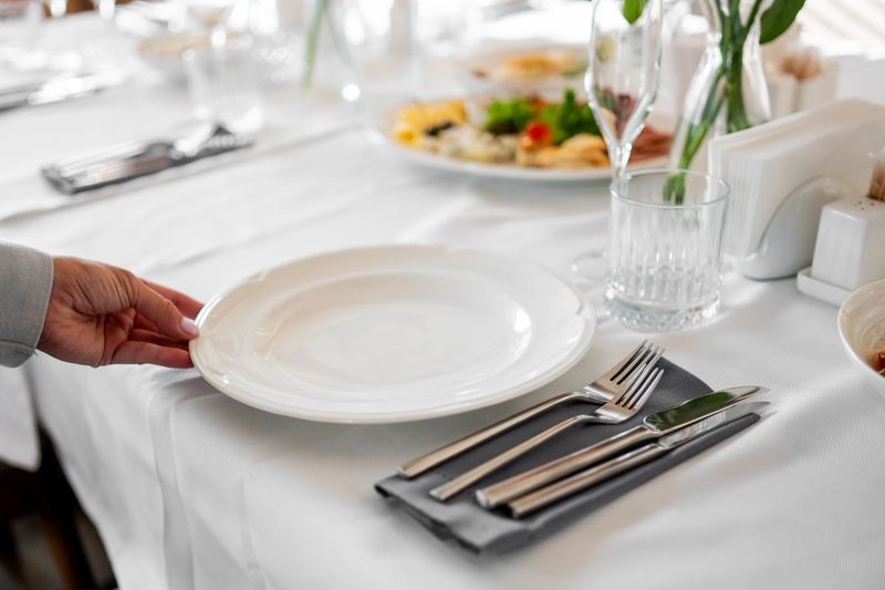 Hand placing a white plate on a neatly set dining table with cutlery, glass, and food in the background, suggesting preparation for a formal meal or event.