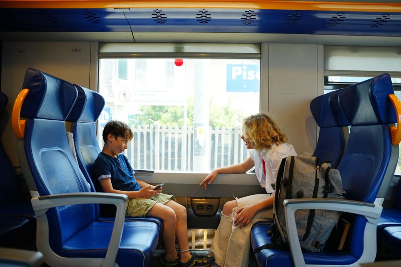 Two children engage in a lively conversation on a train journey, sharing a moment of connection while seated across from each other. The scene captures a sense of curiosity and companionship during travel.