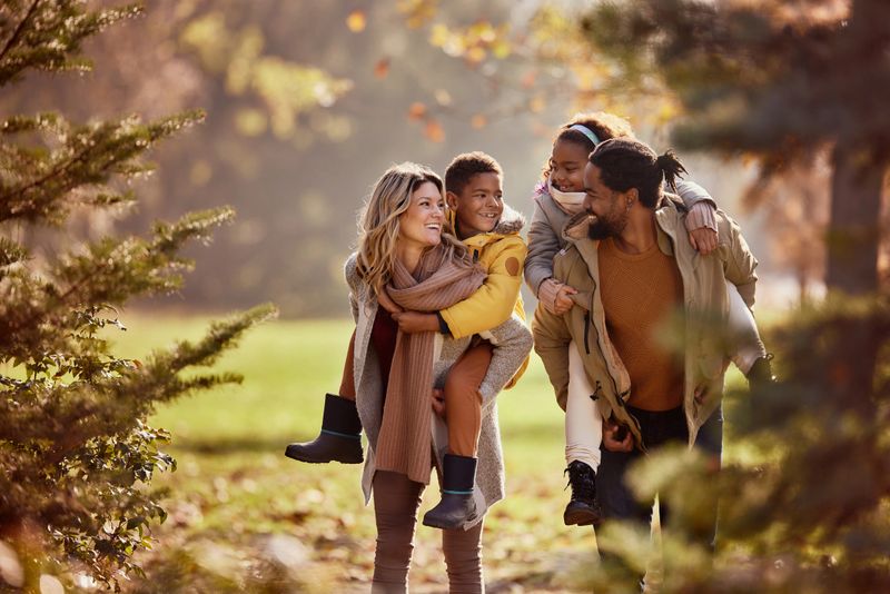 Happy diverse parents having fun while piggybacking their small kids during autumn day in nature.