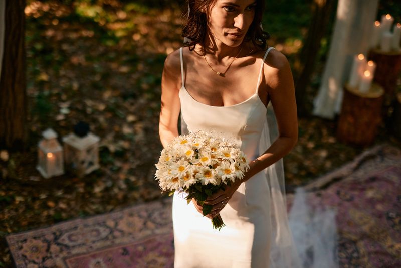 Elegant bride in a white wedding dress holding a bouquet of daisies during a forest wedding ceremony, creating a romantic and natural atmosphere
