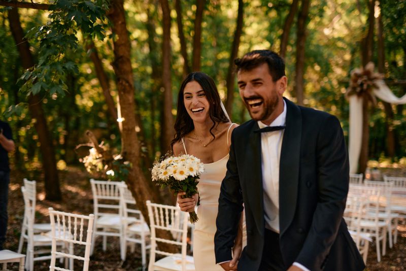 Bride and groom walking hand in hand away from the altar, sharing laughter and joy, surrounded by lush trees and elegant white chairs, celebrating their love in a beautiful outdoor setting