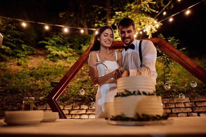 Newlywed couple joyfully cutting their wedding cake during a romantic evening reception, celebrating love and commitment in a beautifully decorated outdoor setting