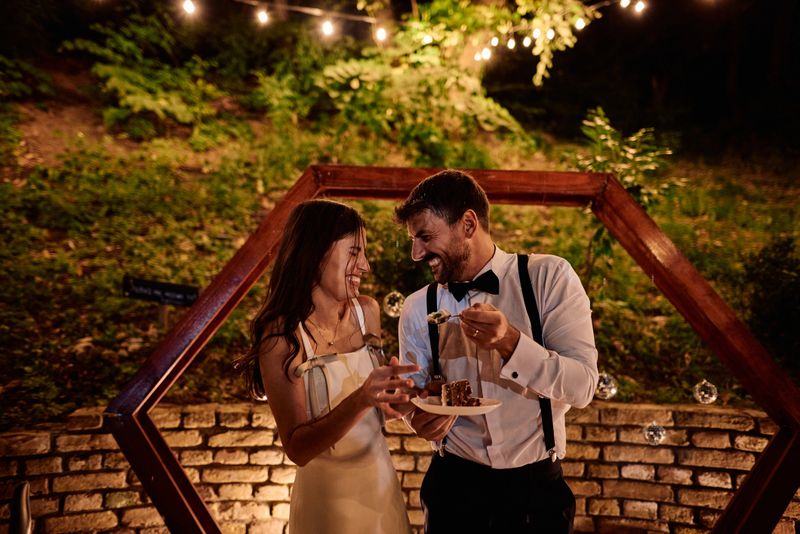 Bride and groom laughing and enjoying cake together under twinkling string lights at their enchanting outdoor wedding reception, celebrating love and togetherness in a joyful evening garden setting