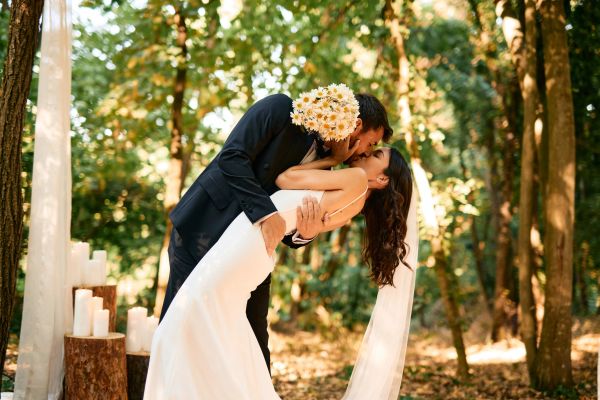 Newlyweds share a romantic kiss in a forest setting.