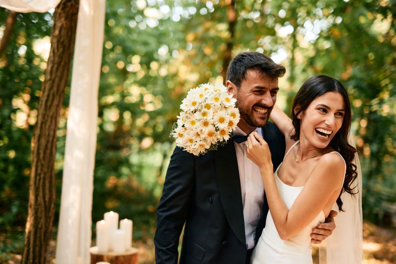 Young bride and groom are laughing and hugging during their wedding ceremony in the woods, with the bride playfully holding a bouquet of daisies near the groom's face