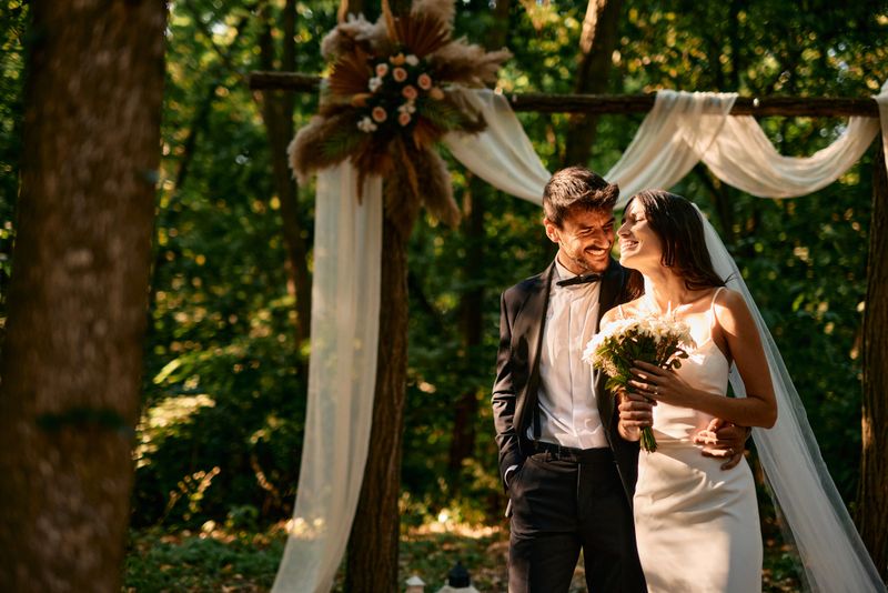 Young bride and groom are standing together in a forest, smiling at each other during their wedding ceremony, surrounded by trees and decorated with white fabrics and flowers