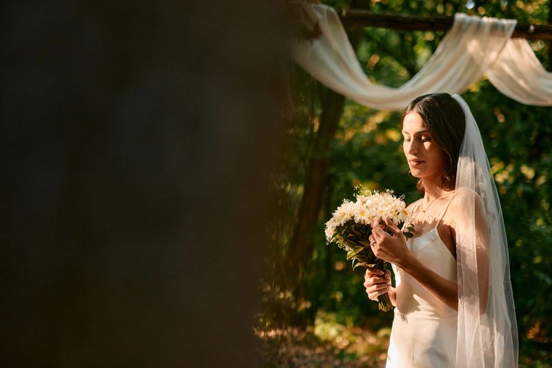 Gorgeous bride holding a bouquet of vibrant daisies poses gracefully in a serene forest, framed by a beautiful wedding arch while wearing an elegant, simple white dress