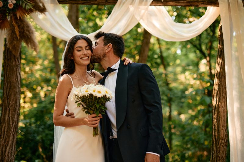 Happy newlywed couple sharing a tender kiss beneath a beautifully decorated wooden arch in a serene forest, celebrating their love and commitment in a romantic outdoor setting