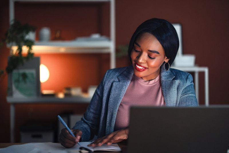 A confident woman in a modern home office space, surrounded by shelves and warm lighting, writing notes energetically while sitting at her desk, projecting professionalism and productivity.