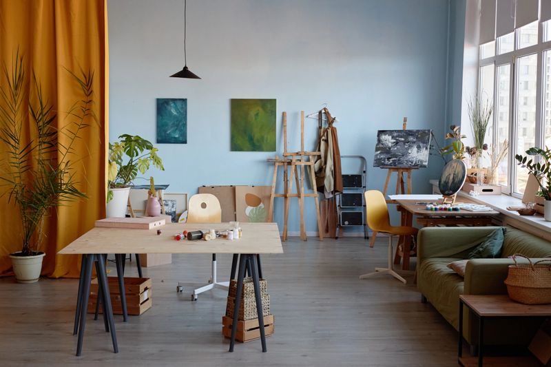 Spacious art studio featuring large wooden tables, easels with canvases, various art supplies and potted plants arranged near tall windows providing abundant natural light