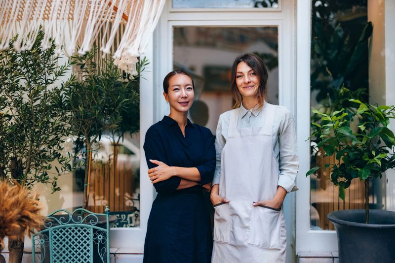 Two women standing together near a storefront, surrounded by lush green plants, symbolizing collaboration, entrepreneurship, and urban lifestyle.