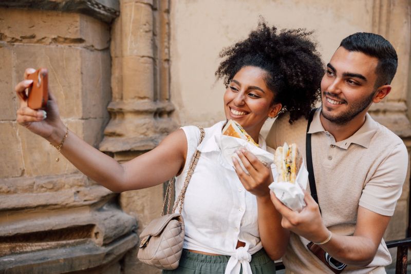 A cheerful couple stands outside a historical building in Italy, sharing a moment of joy while enjoying delicious street food. The woman, wearing a white blouse and green pants, smiles as she holds her food, while the man, dressed in a beige polo shirt and white pants, also enjoys his meal. The warm sun, casual outfits, and the rustic architecture create a relaxed, vacation vibe.