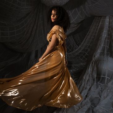Woman in a flowing golden dress poses against a dark, draped backdrop.