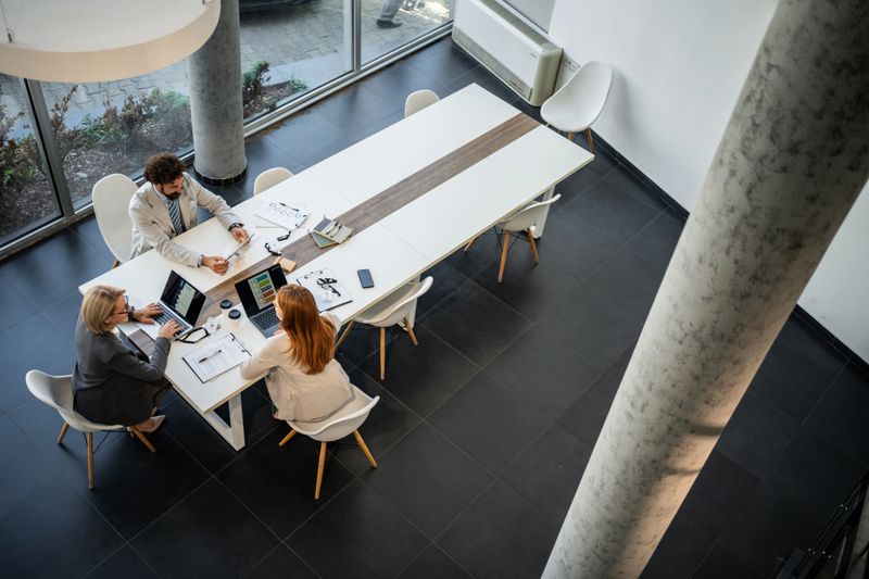 Three businesspeople are sitting at a large white table in a modern office, collaborating on a project using laptops and documents, seen from a high angle view