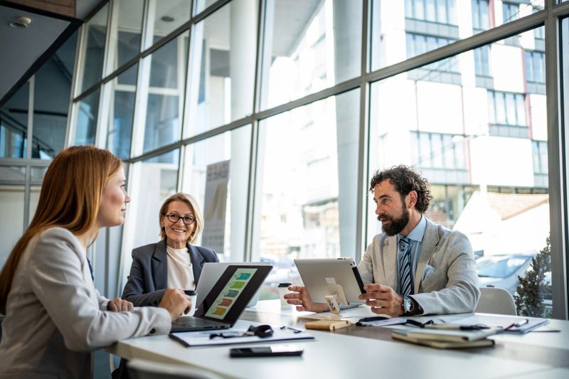 Three business people are sitting at a table in a modern office, using a digital tablet and laptop to discuss a project, smiling and collaborating effectively
